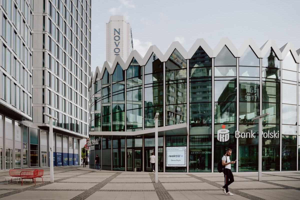 fotograf startupow rotunda reaktorX 4 Ein modernes Glasgebäude mit einem Zickzackdach, auf dem das Logo der 'Bank of Poland' zu sehen ist. Draußen geht eine Person vorbei und eine andere sitzt auf einer roten Bank. Im Hintergrund sind hohe Gebäude zu sehen, darunter eines mit der Aufschrift 'Novotel'.