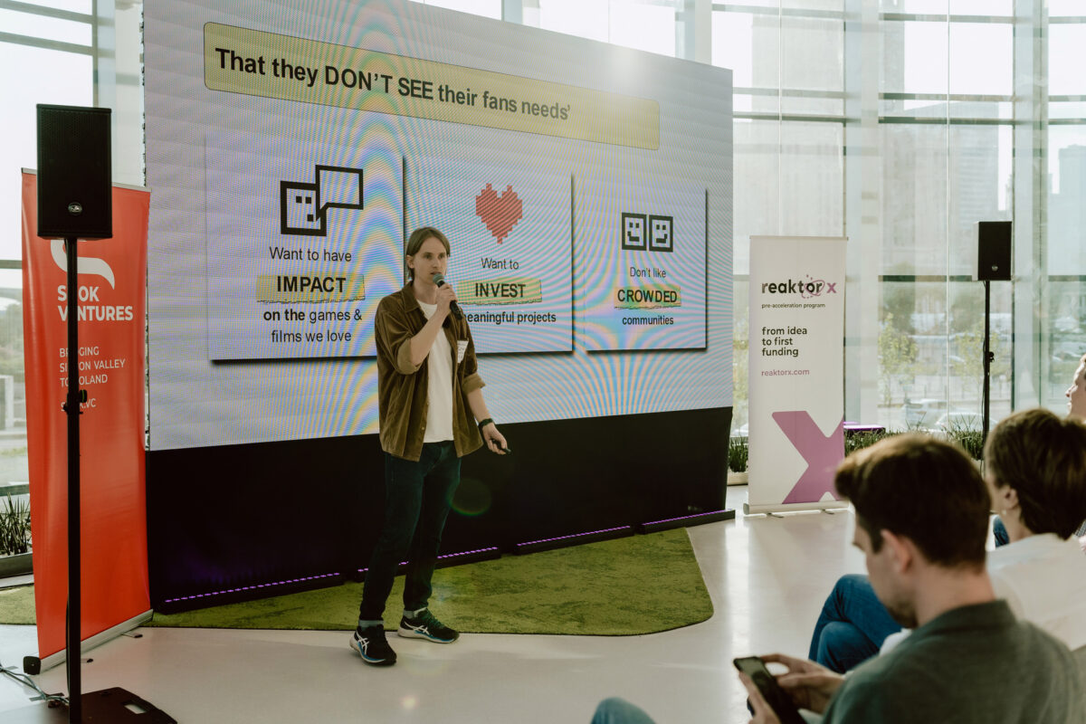 fotograf startupow rotunda reaktorX 40 Ein Mann spricht in ein Mikrofon auf der Bühne vor einem großen Bildschirm, auf dem Diagramme und Text angezeigt werden. Im Publikum sitzen Menschen. Auf beiden Seiten sind Banner mit den Aufschriften 'Sisu Ventures' und 'Reaktor X' angebracht.