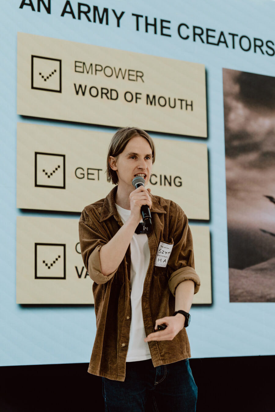 fotograf startupow rotunda reaktorX 41 Eine Person mit langen braunen Haaren, die eine braune Jacke trägt und ein Mikrofon in der Hand hält, spricht vor einer Präsentationsfolie mit den Worten 'EMPOWER WORD OF MOUTH, GET PRICING, CREATE VALUE'.