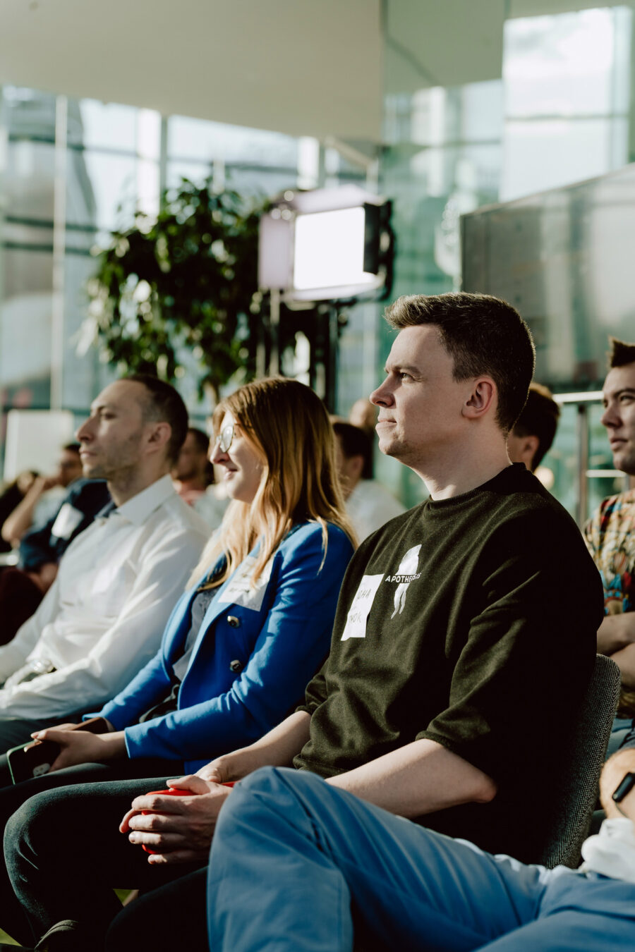 fotograf startupow rotunda reaktorX 42 Eine Gruppe von Menschen sitzt in einem Raum in Reihen und beobachtet aufmerksam etwas außerhalb des Rahmens. Das Sonnenlicht fällt durch große Fenster ein, und im Hintergrund sind eine Topfpflanze und eine Studioleuchte zu sehen.