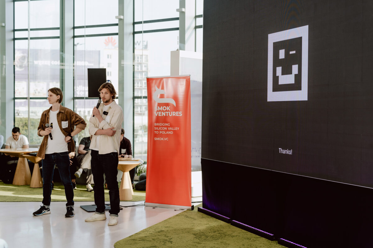 fotograf startupow rotunda reaktorX 43 Zwei Männer stehen mit Mikrofonen auf der Bühne. Ein großer Bildschirm zeigt ein verpixeltes Smiley und die Worte "Danke!". Neben ihnen ist ein rotes SMOK Ventures-Banner zu sehen. Die Leute sitzen an Tischen in einem modernen, hellen Raum.