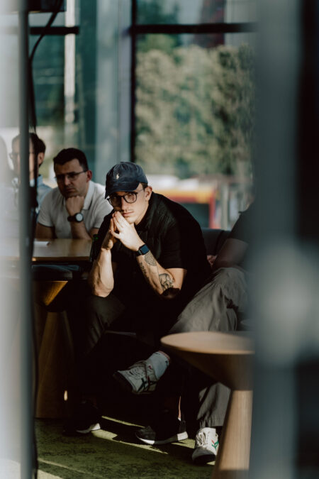 fotograf startupow rotunda reaktorX 45 Ein Mann mit Mütze, Brille und dunkler Kleidung sitzt nachdenklich an einem Tisch mit anderen und stützt sein Kinn in einem hellen Raum mit großen Fenstern auf seine Hände.