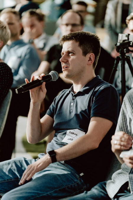 fotograf startupow rotunda reaktorX 46 Ein Mann in einem marineblauen Polohemd sitzt auf einer Veranstaltung und spricht in ein Mikrofon, er trägt ein Namensschild. Um ihn herum sitzen andere Teilnehmer und im Hintergrund ist eine Kamera auf einem Stativ zu sehen.