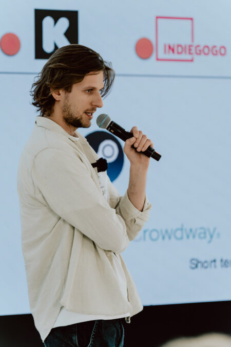 fotograf startupow rotunda reaktorX 47 Ein Mann mit einem Mikrofon spricht auf der Bühne vor einem Bildschirm, auf dem die Logos von Kickstarter, Indiegogo und anderen Crowdfunding-Plattformen zu sehen sind.