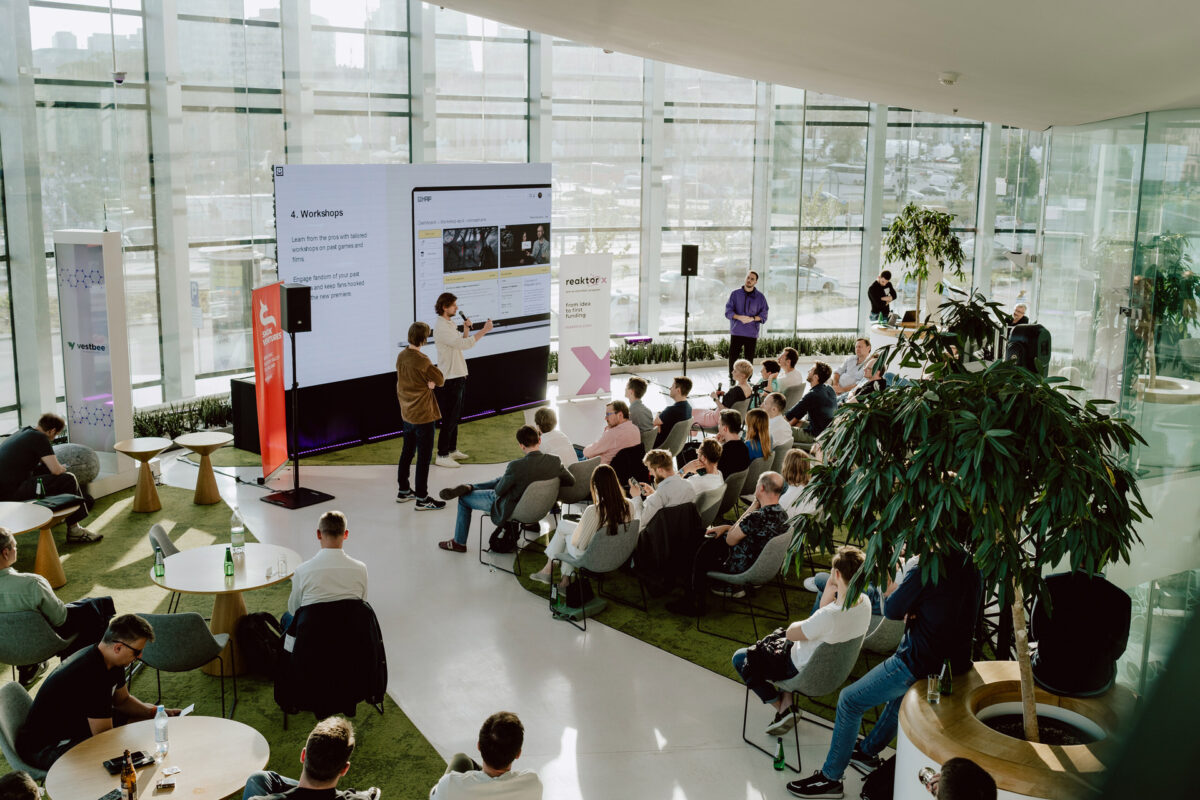 fotograf startupow rotunda reaktorX 48 Eine Gruppe von Personen sitzt in einem modernen, verglasten Konferenzraum mit Pflanzen und Blick auf die Stadt und hört zwei Rednern zu, die vor einem großen Bildschirm präsentieren.