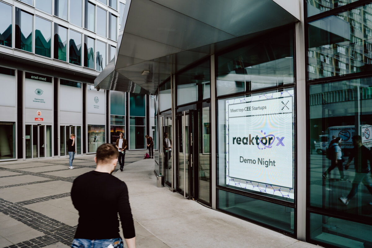fotograf startupow rotunda reaktorX 5 Eine Person geht an einem modernen Glasgebäude mit einem großen digitalen Schild "Meet top CEE Startups, reactorX Demo Night" in der Nähe des Eingangs vorbei. Im Hintergrund sind weitere Personen zu sehen.