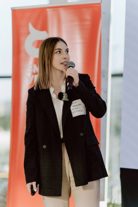 fotograf startupow rotunda reaktorX 52 Eine Frau in einer schwarzen Jacke und mit einem Mikrofon in der Hand steht vor einem roten Banner und spricht oder präsentiert. Sie trägt ein Namensschild mit der Aufschrift 'Marta' und schaut zur Seite.