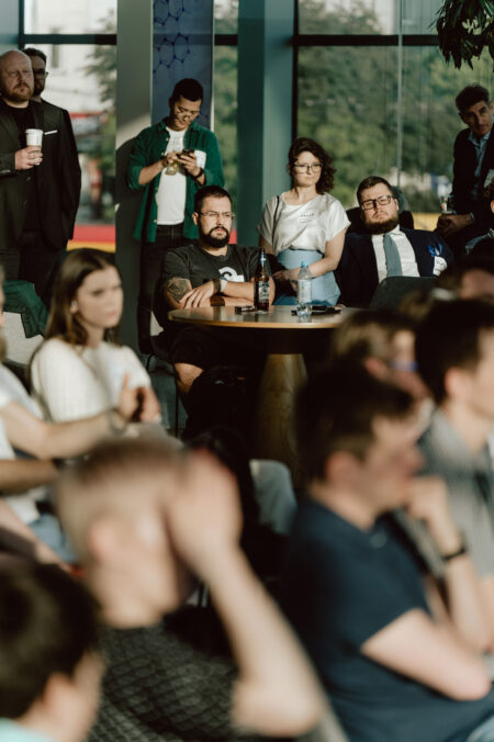 fotograf startupow rotunda reaktorX 53 Eine Gruppe von Menschen sitzt und steht in einem Raum und beobachtet aufmerksam eine Veranstaltung oder Präsentation. Einige halten Getränke in der Hand. Die Bühne wird von natürlichem Sonnenlicht beleuchtet, das durch große Fenster fällt. Die Stimmung scheint konzentriert und engagiert zu sein.
