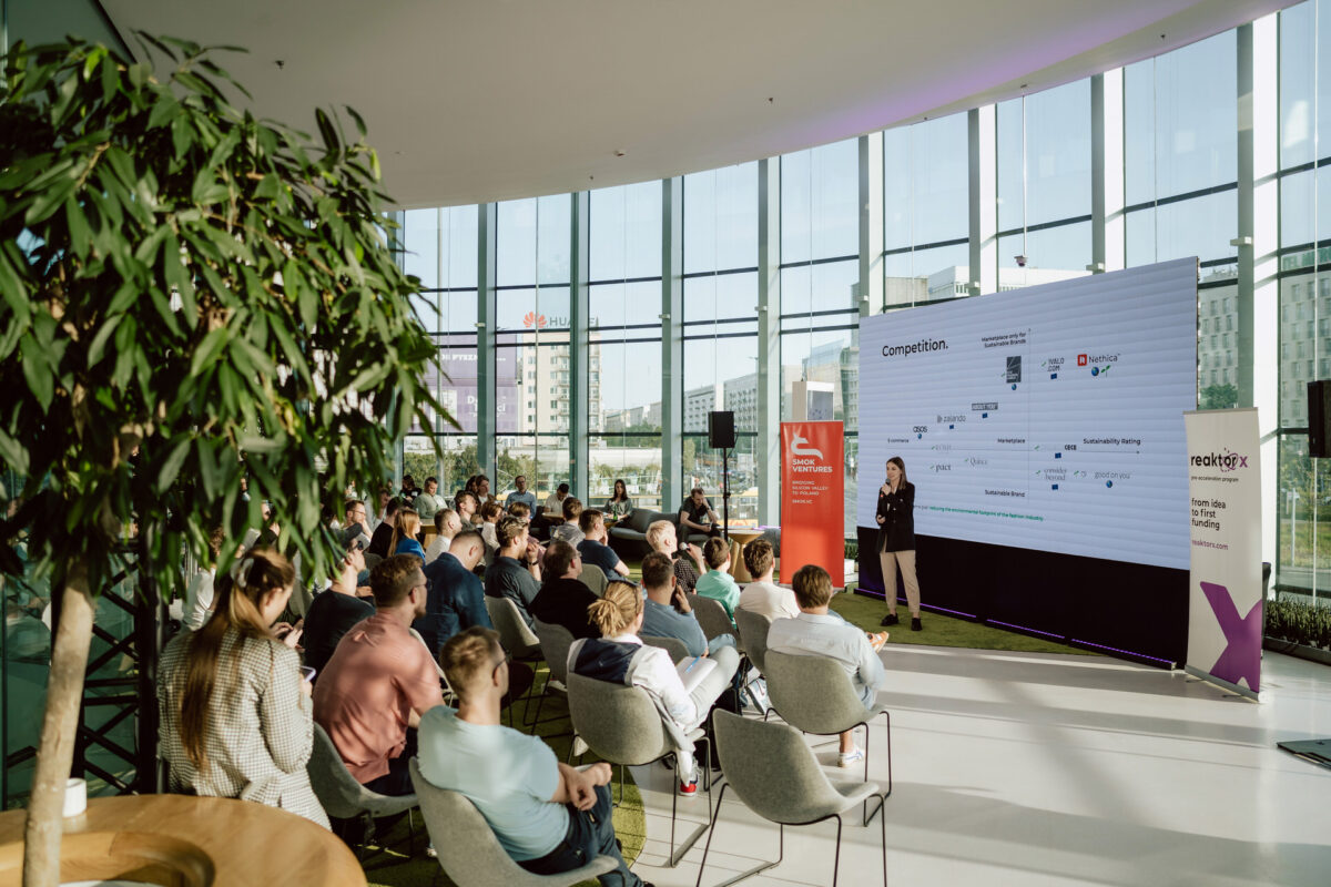 fotograf startupow rotunda reaktorX 54 Der Redner spricht vor einem großen Bildschirm vor einem Publikum, das in einem modernen, hellen Raum mit raumhohen Fenstern und Blick auf die Stadt sitzt. Auf der linken Seite des Bildes ist eine große Pflanze zu sehen.