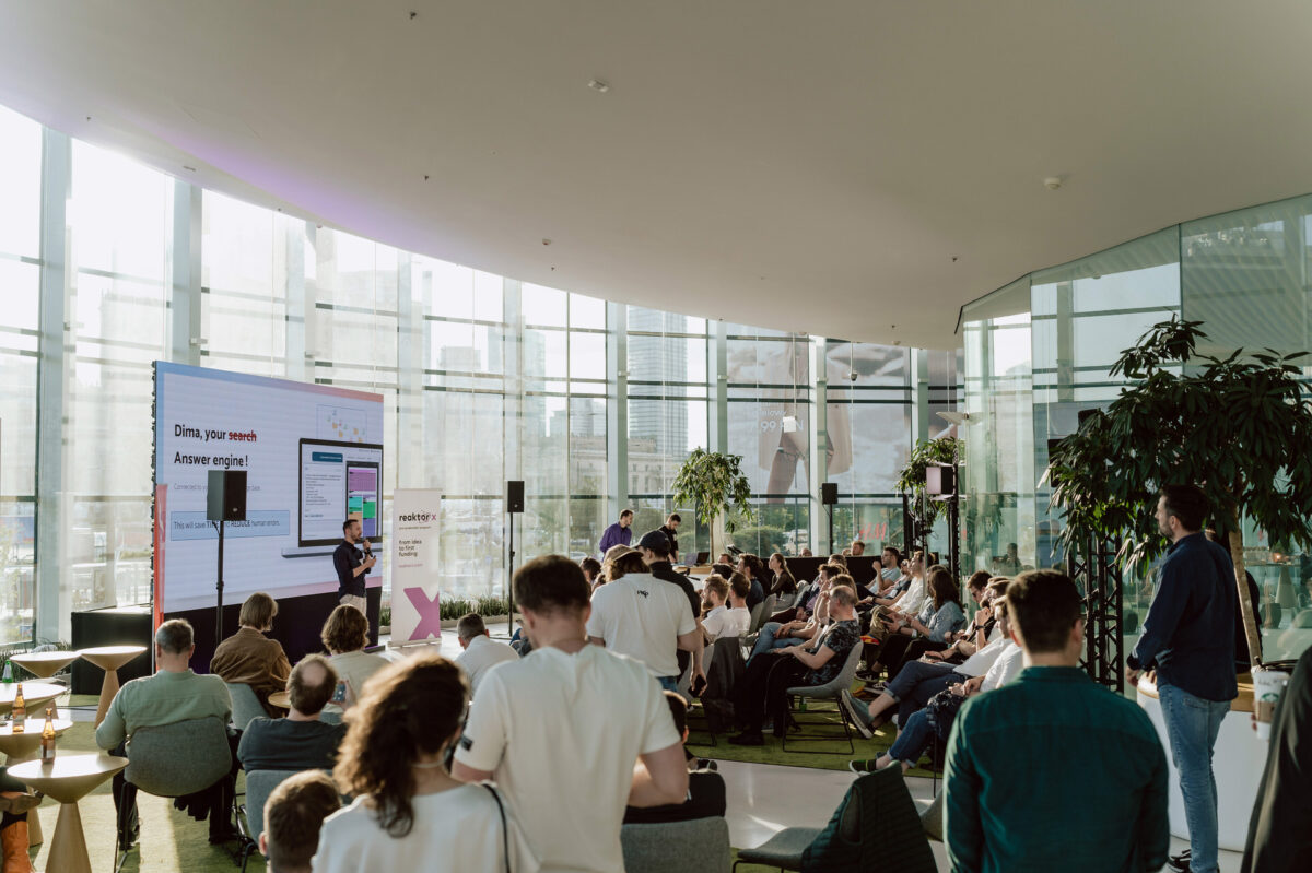 fotograf startupow rotunda reaktorX 56 Der Redner spricht vor einem Publikum in einem modernen, sonnendurchfluteten Konferenzraum mit bodentiefen Fenstern, großen Bildschirmen und Menschen, die aufmerksam sitzen und zuhören.