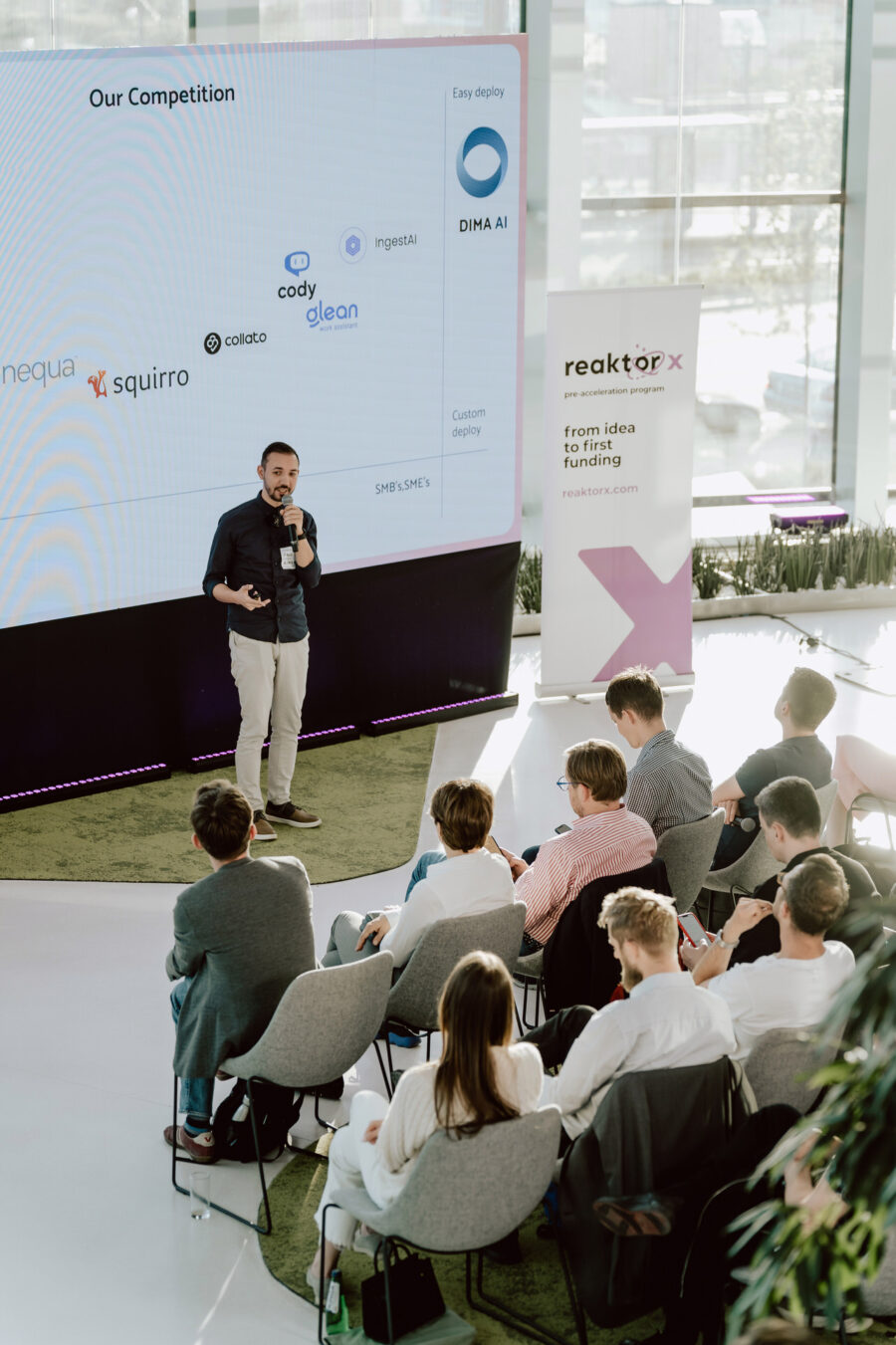 fotograf startupow rotunda reaktorX 57 Ein Mann steht auf der Bühne und hält eine Präsentation vor einem sitzenden Publikum. Hinter ihm befindet sich ein großer Bildschirm, auf dem ein Diagramm zur Wettbewerbsanalyse und ein Logo zu sehen sind, während an der Seite ein Banner mit der Marke 'reactorx' zu sehen ist.