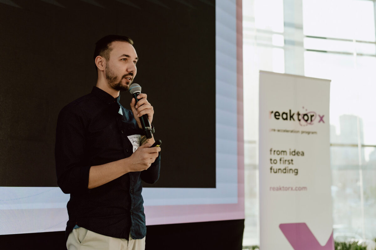 fotograf startupow rotunda reaktorX 59 Ein Mann mit einem Mikrofon spricht auf der Bühne neben einem großen Bildschirm. Hinter ihm steht ein Banner mit der Aufschrift 'Reaktor X: von der Idee zur ersten Finanzierung'. Durch die großen Fenster im Hintergrund fällt Sonnenlicht ein.