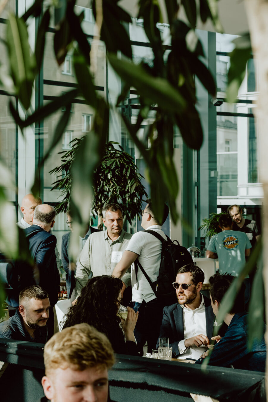 fotograf startupow rotunda reaktorX 6 Eine Gruppe von Menschen trifft sich an einem sonnenbeschienenen Ort mit großen Fenstern und üppiger Bepflanzung. Einige sitzen an Tischen und unterhalten sich, während andere im Hintergrund stehen und sich unter die Leute mischen.