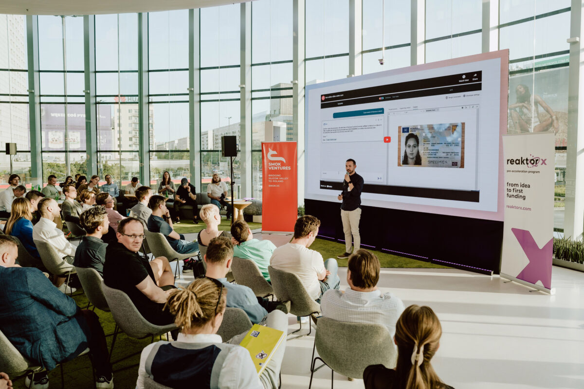 fotograf startupow rotunda reaktorX 60 Der Redner spricht vor einem Publikum in einem modernen, sonnendurchfluteten Raum mit großen Fenstern. Ein großer Bildschirm hinter dem Redner zeigt eine Website-Oberfläche an. Die Leute sitzen und hören aufmerksam zu, einige machen sich Notizen. Der Bühnenbereich ist mit Bannern geschmückt.