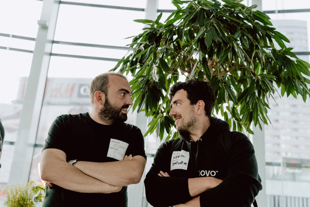 fotograf startupow rotunda reaktorX 62 Zwei Männer stehen mit verschränkten Armen in einem Raum, schauen sich an und lächeln. Sie tragen beide Namensschilder und legere Kleidung. Hinter ihnen steht eine große Blattpflanze und die hellen Fenster zeigen die Gebäude der Stadt draußen.