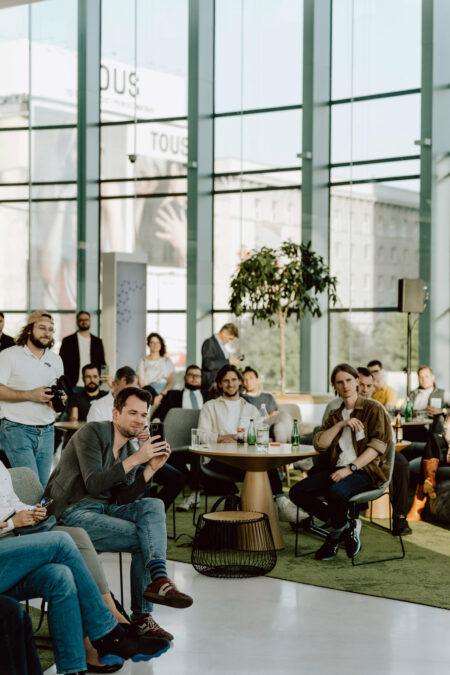 fotograf startupow rotunda reaktorX 64 Eine Gruppe von Menschen sitzt während einer Party in einem modernen, sonnigen Raum mit großen Fenstern und hört aufmerksam zu. Einige halten Getränke in der Hand, während ein Mann im Vordergrund auf sein Telefon schaut.