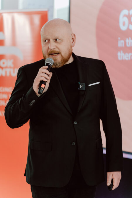 fotograf startupow rotunda reaktorX 65 Ein glatzköpfiger Mann mit Bart, der einen schwarzen Anzug trägt und ein Mikrofon in der Hand hält, spricht bei einer Veranstaltung. Er steht vor einem roten und rosa Präsentationsbildschirm und einem Banner mit teilweise sichtbarem Text.