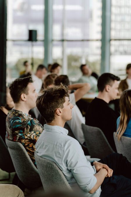 fotograf startupow rotunda reaktorX 67 Eine Gruppe von Menschen sitzt bei einer Veranstaltung oder einem Seminar in einem Raum aufmerksam in Reihen von modernen Stühlen und hört dem Redner außerhalb des Rahmens zu. Die Umgebung ist hell mit großen Fenstern im Hintergrund.