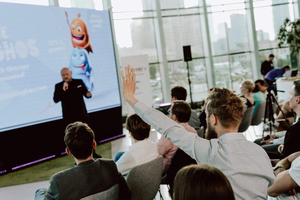 fotograf startupow rotunda reaktorX 68 Ein Mann aus dem Publikum hebt die Hand, um während einer Präsentation in einem modernen, hellen Konferenzraum eine Frage zu stellen. Der Redner steht neben einem Bildschirm, auf dem animierte Figuren und Text zu sehen sind.