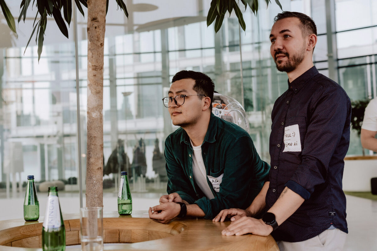 fotograf startupow rotunda reaktorX 69 Zwei Männer stehen an einem runden Holztisch und schauen aufmerksam nach vorne. Sie tragen beide Namensschilder und einfache Hemden. Auf dem Tisch stehen grüne Glaswasserflaschen und im Hintergrund sind große Fenster mit weichem Licht zu sehen.