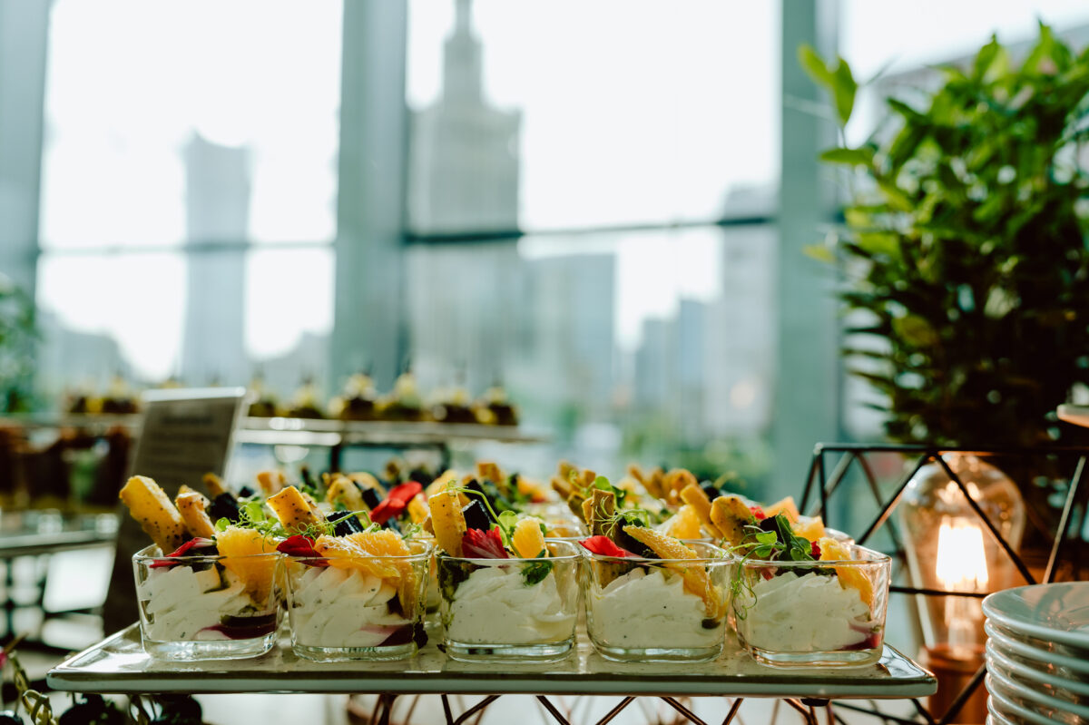 fotograf startupow rotunda reaktorX 7 Eine Reihe von Glasbechern, gefüllt mit geschichteten Snacks, darunter Sahne, Grünzeug und geröstete Brotscheiben, die auf einem Tablett präsentiert werden, während die Skyline der Stadt durch die großen Fenster im Hintergrund zu sehen ist.