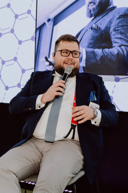 fotograf startupow rotunda reaktorX 70 Ein Mann in Anzug und Krawatte sitzt während der Veranstaltung und spricht in ein Mikrofon, während ein großer Bildschirm hinter ihm sein Bild und das Muster des Netzwerks anzeigt.