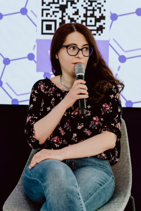 fotograf startupow rotunda reaktorX 71 Eine Frau mit langen braunen Haaren und Brille, die ein schwarzes geblümtes Oberteil und Jeans trägt, sitzt auf einem grauen Stuhl und hält ein Mikrofon. Im Hintergrund ist ein großer Bildschirm mit einem QR-Code und einem abstrakten Muster zu sehen.