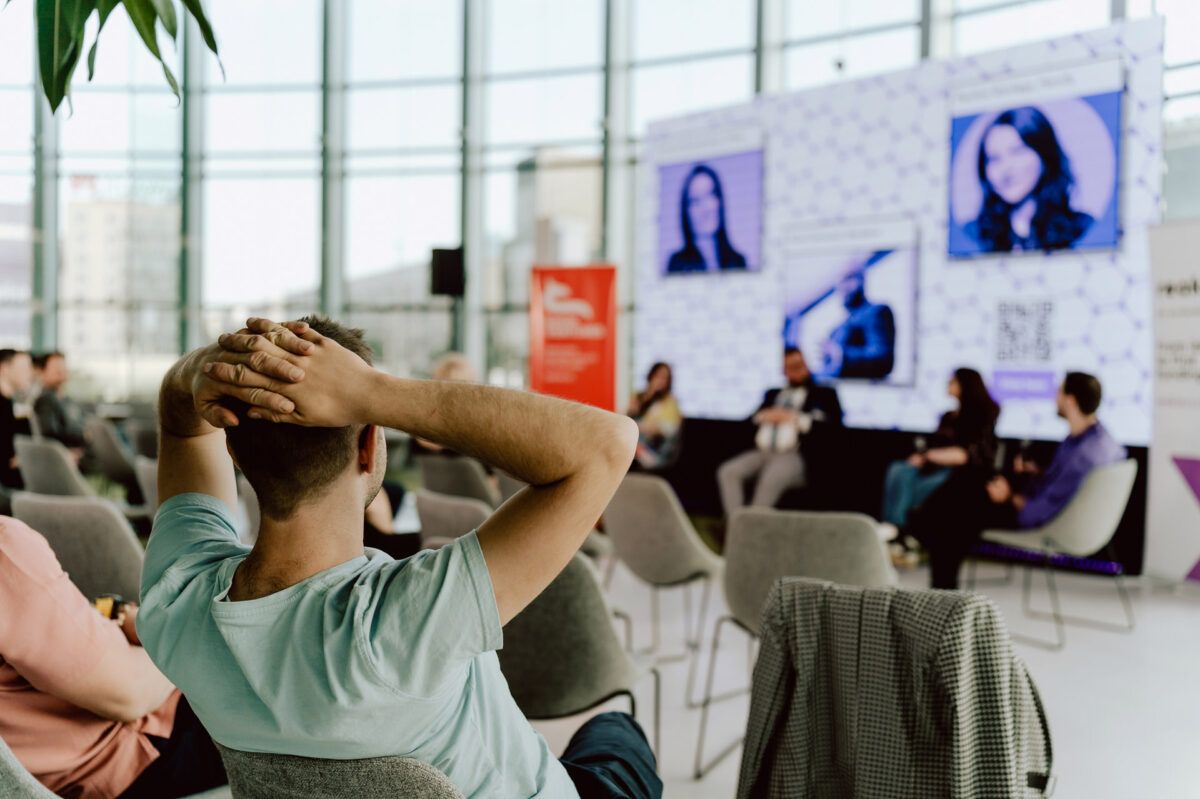 fotograf startupow rotunda reaktorX 72 Eine Person sitzt mit den Händen auf dem Kopf vor einer Podiumsdiskussion in einem modernen Raum mit Glaswänden. Mehrere Personen sitzen im Publikum, während die Redner auf der Bühne mit digitalen Bildschirmen hinter ihnen erscheinen.