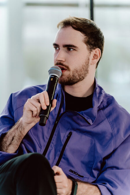 fotograf startupow rotunda reaktorX 73 Ein junger Mann mit kurzen braunen Haaren und Bart, der eine lila Jacke trägt, hält ein Mikrofon und spricht. Er blickt leicht nach links in den gut beleuchteten Raum.