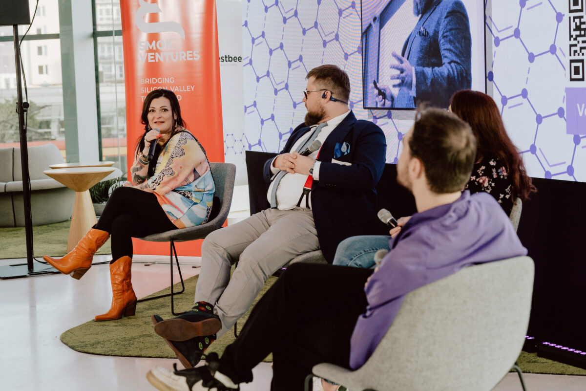 fotograf startupow rotunda reaktorX 74 Vier Personen sitzen während einer Podiumsdiskussion auf der Bühne. Eine Frau auf der linken Seite, die ein buntes Outfit und orangefarbene Schuhe trägt, spricht in ein Mikrofon. Zwei Männer und eine weitere Frau hören zu. Hinter ihnen befinden sich ein großer Bildschirm und ein Veranstaltungsbanner.