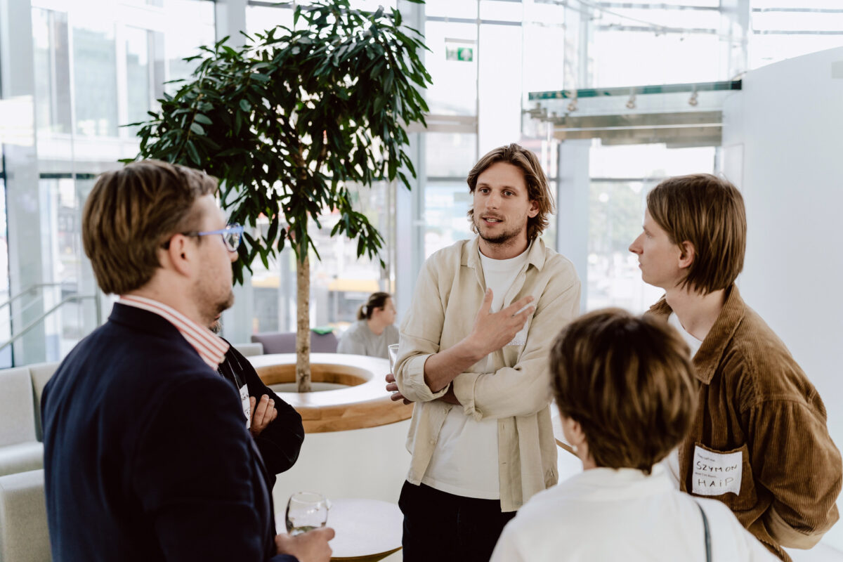 fotograf startupow rotunda reaktorX 75 Vier Personen stehen in einem modernen, hellen Raum und führen ein Gespräch. Eine Person gestikuliert mit ihren Händen, während die anderen aufmerksam zuhören. Im Hintergrund stehen eine große Topfpflanze und Glaswände.