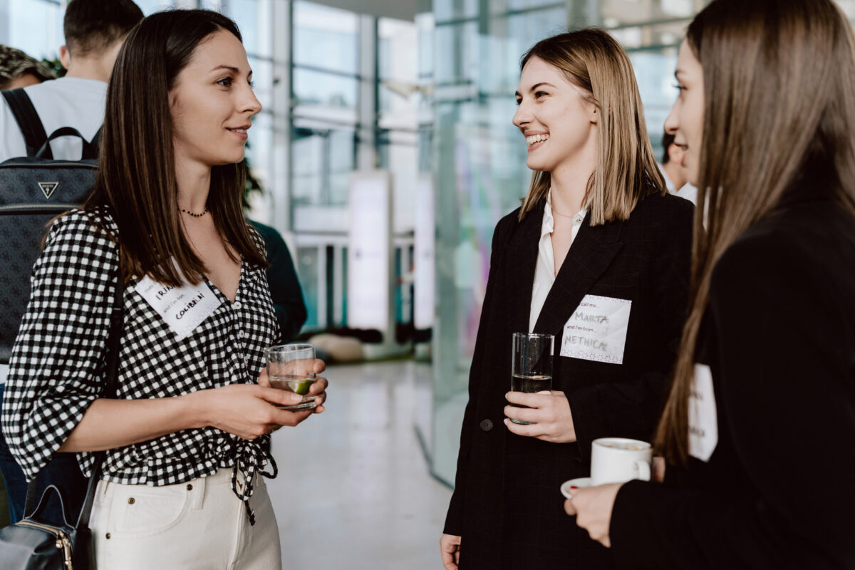 fotograf startupow rotunda reaktorX 76 Drei Frauen stehen bei einem Networking-Treffen in einem Raum, tragen Namensschilder und halten Getränke in der Hand. Sie lächeln und unterhalten sich in einem hellen, modernen Raum mit Glaswänden.