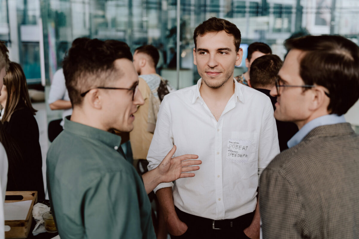fotograf startupow rotunda reaktorX 77 Drei Männer unterhalten sich bei einem gesellschaftlichen Treffen oder einer Netzwerkveranstaltung. Ein Mann in einem weißen Hemd mit einem Abzeichen steht in der Mitte, während die beiden anderen ihm gegenüber stehen und sich unterhalten. Im Hintergrund sind weitere Personen zu sehen.