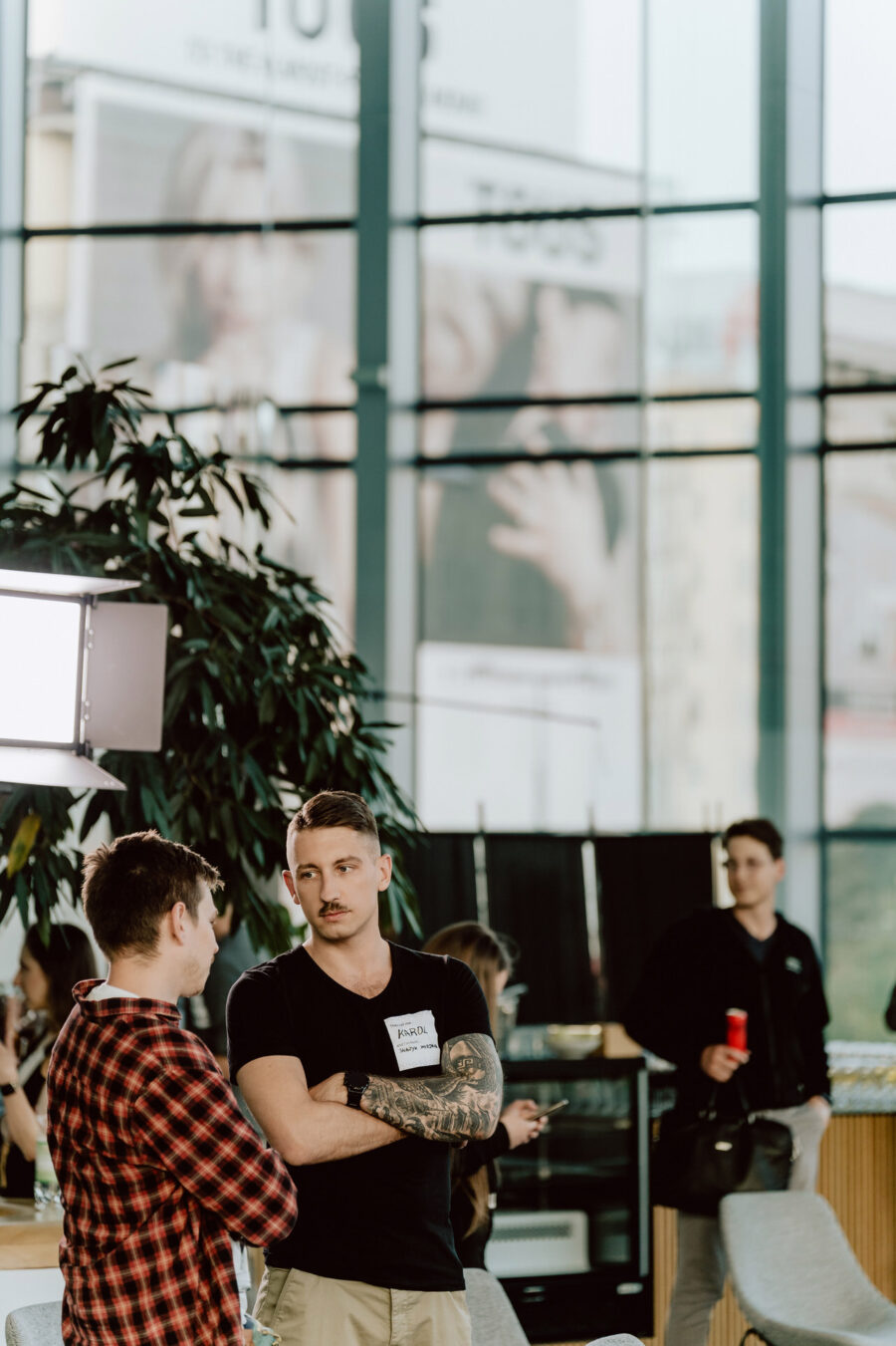 fotograf startupow rotunda reaktorX 78 Zwei Männer stehen und unterhalten sich in einem Raum in der Nähe eines großen Fensters. Der eine trägt ein rotkariertes Hemd, der andere ein schwarzes T-Shirt mit Tattoos und einem Namensschild. Im Hintergrund sind andere Personen und eine Pflanze zu sehen.