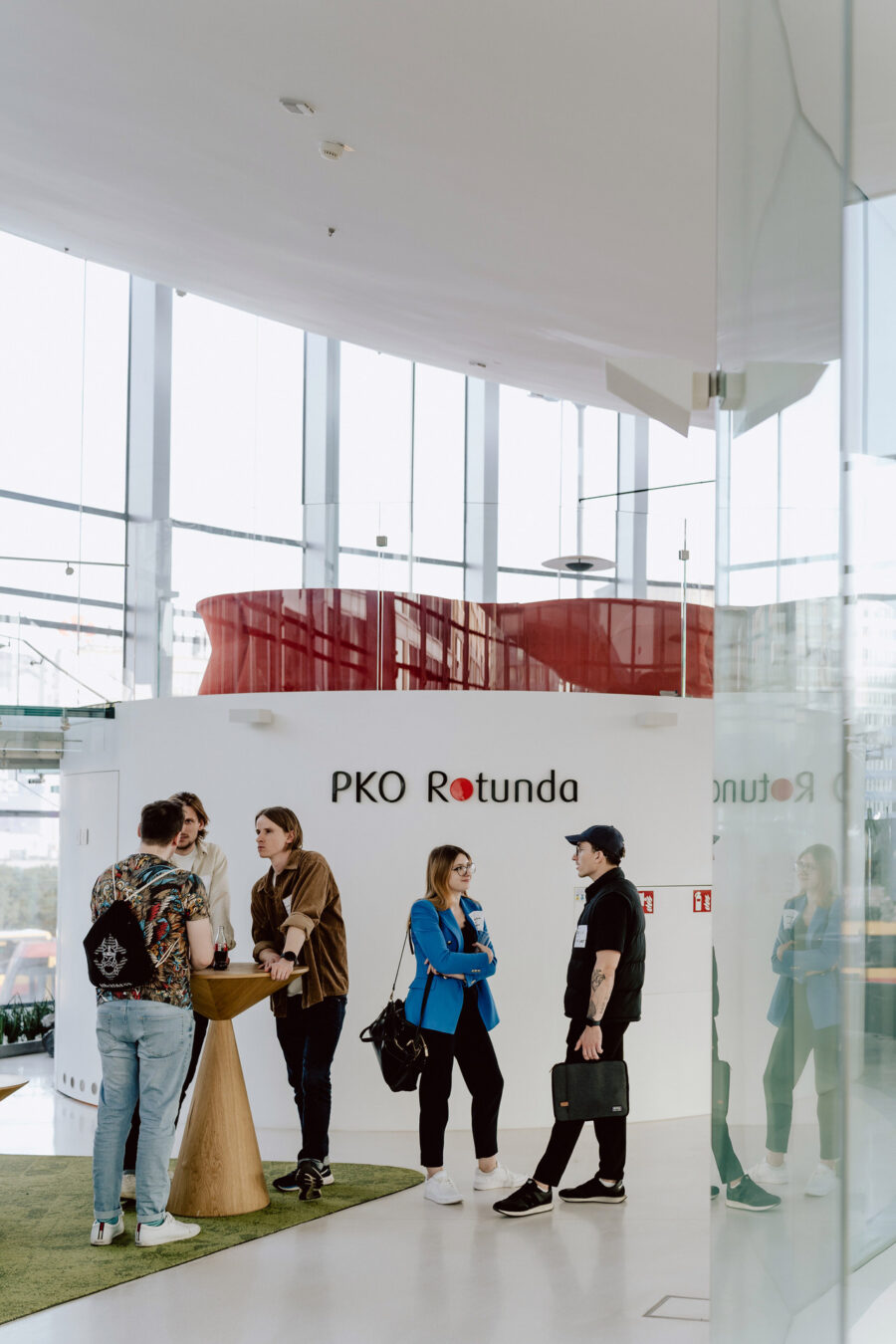 fotograf startupow rotunda reaktorX 79 Eine Gruppe von fünf jungen Leuten unterhält sich an einem Holztisch in einer hellen, modernen Lobby mit großen Fenstern. An der Wand hinter ihnen prangt das Logo 'PKO Rotunda'.