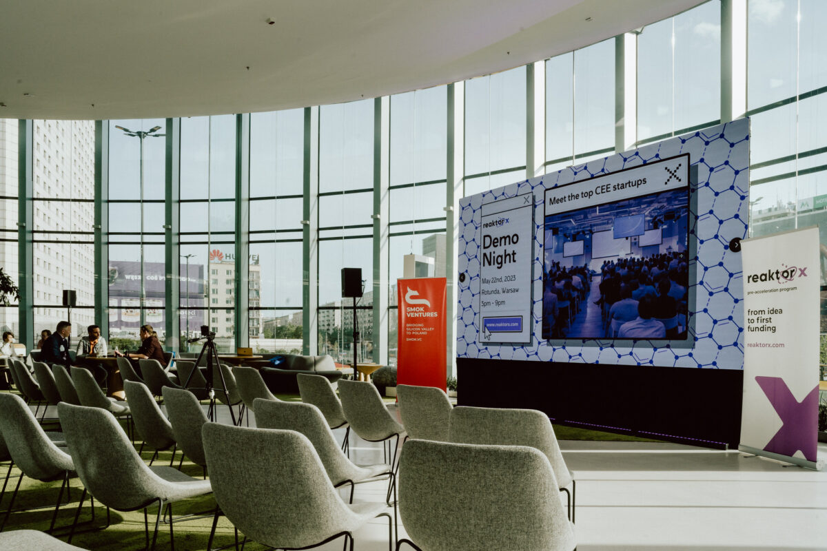 fotograf startupow rotunda reaktorX 8 Ein moderner Konferenzraum mit Reihen leerer Stühle gegenüber einem großen Bildschirm, auf dem 'Demo Night' und Grafiken zum Thema Startup zu sehen sind. Auf der linken Seite sitzen nur wenige Leute, und die hohen Fenster zeigen die Gebäude der Stadt draußen.