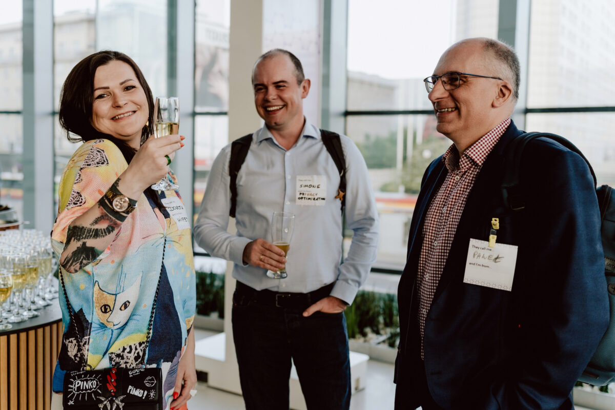 fotograf startupow rotunda reaktorX 80 Drei lächelnde Menschen stehen bei einem Networking-Event in einem Raum an großen Fenstern, halten Getränke in der Hand und tragen Namensschilder. Eine Frau erhebt ein Glas und zwei Männer stehen neben ihr, alle sehen fröhlich und engagiert aus.