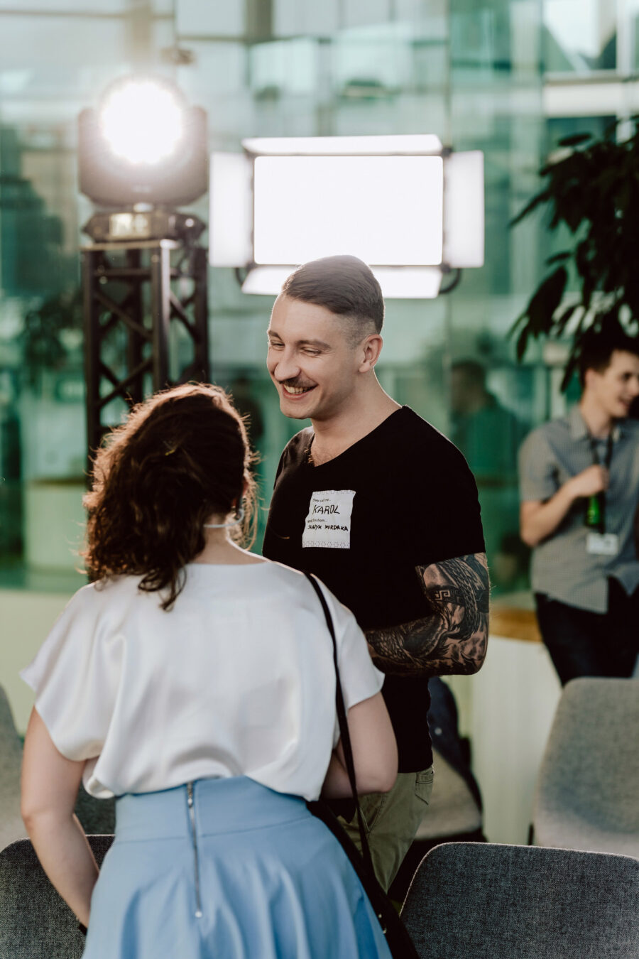 fotograf startupow rotunda reaktorX 82 Ein Mann mit Tätowierungen und einem Namensschild lächelt, während er sich mit einer Frau mit lockigem Haar auf einer Zimmerparty unterhält. Helle Studiolichter und eine weitere Person im Hintergrund sind zu sehen.