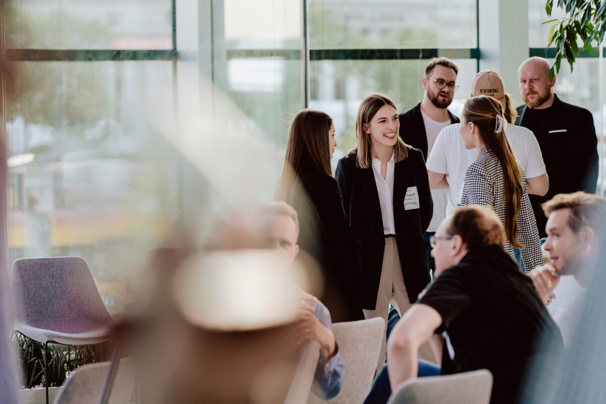fotograf startupow rotunda reaktorX 83 Eine Gruppe von Menschen plaudert und mischt sich in einem hellen, modernen Raum mit großen Fenstern. Einige stehen und reden, während andere sitzen und diskutieren.