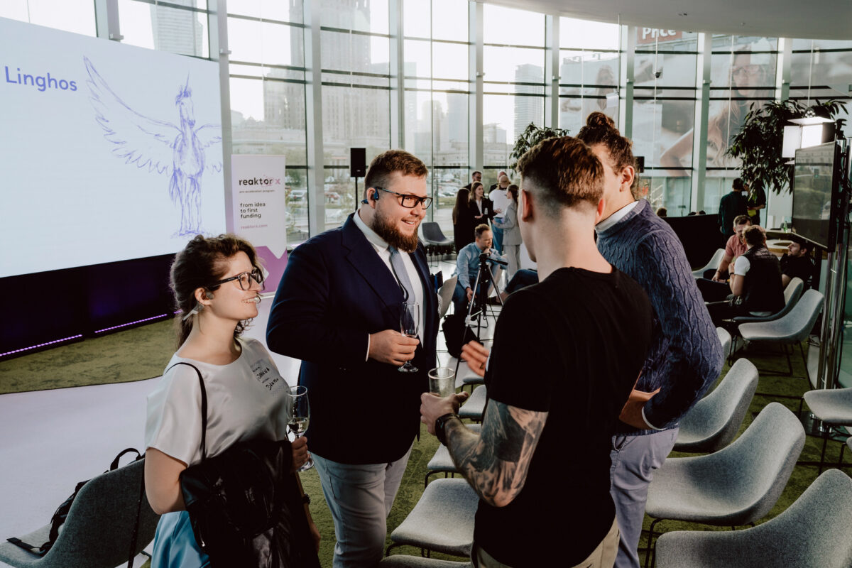 fotograf startupow rotunda reaktorX 84 Vier Personen stehen im Kreis, unterhalten sich und halten Getränke in einem modernen Interieur mit großen Fenstern und Blick auf die Stadt. Der Bildschirm hinter ihnen zeigt die Illustration eines blauen Vogels und Text. Andere Teilnehmer sitzen in der Nähe.