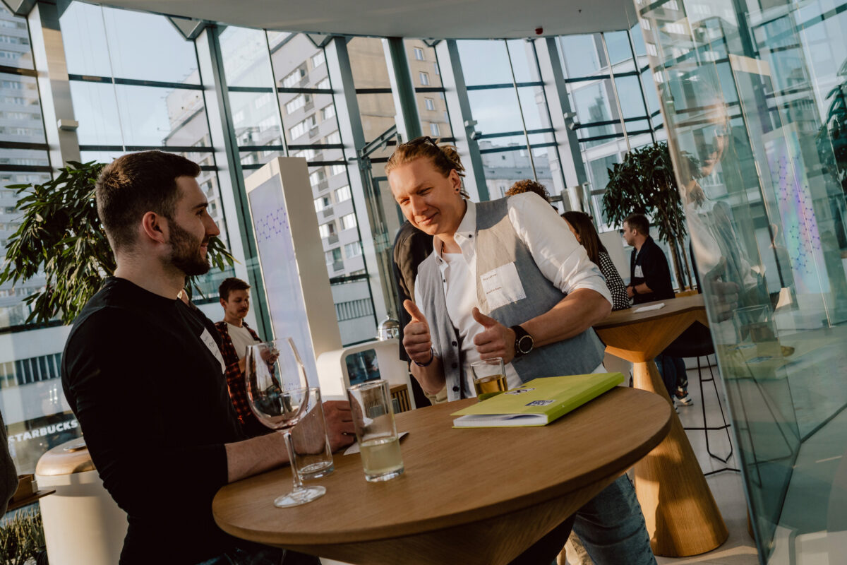 fotograf startupow rotunda reaktorX 85 Ein Gespräch zwischen zwei Männern, die an einem runden Tisch in einem hellen, modernen Raum mit großen Fenstern stehen. Einer der Männer hält den Daumen hoch und lächelt, während die anderen im Hintergrund miteinander sprechen.