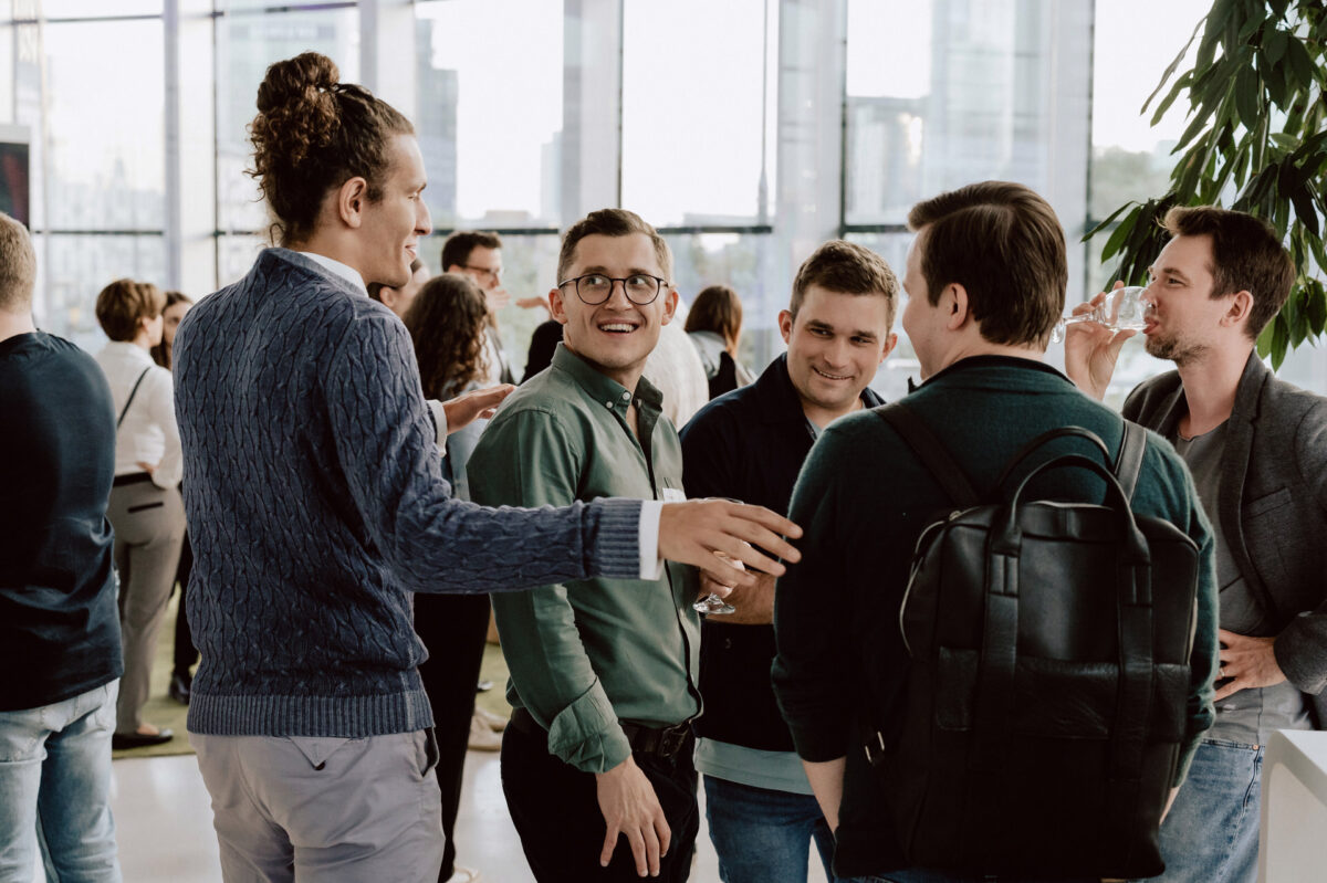 fotograf startupow rotunda reaktorX 89 Eine Gruppe von Menschen steht bei einem gesellschaftlichen Ereignis in einem Raum zusammen, unterhält sich und lächelt. Im Hintergrund sind große Fenster und städtische Gebäude zu sehen. Eine Person trinkt aus einem Glas.