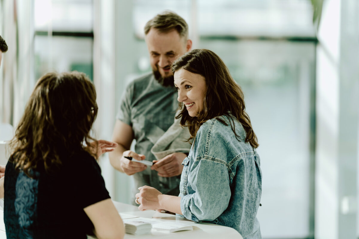 fotograf startupow rotunda reaktorX 9 Drei Erwachsene stehen an einem weißen Tisch und unterhalten sich. Eine Frau in einem Jeanshemd lächelt und hält einen Stift in der Hand, während ein Mann und eine weitere Frau zuhören und sich unterhalten.