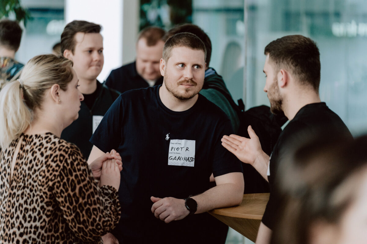 fotograf startupow rotunda reaktorX 90 Eine Gruppe von Menschen unterhält sich an einem Holztisch. Ein Mann, der ein schwarzes T-Shirt mit seinem Namen trägt, hört einem anderen Mann aufmerksam zu, der mit den Händen gestikuliert, während eine Frau und andere Personen in der Nähe ein Gespräch führen.