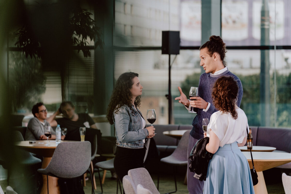 fotograf startupow rotunda reaktorX 91 Drei Personen stehen in einem modernen, gut beleuchteten Café, halten Weingläser in der Hand und führen ein Gespräch, während andere Gäste im Hintergrund sitzen.