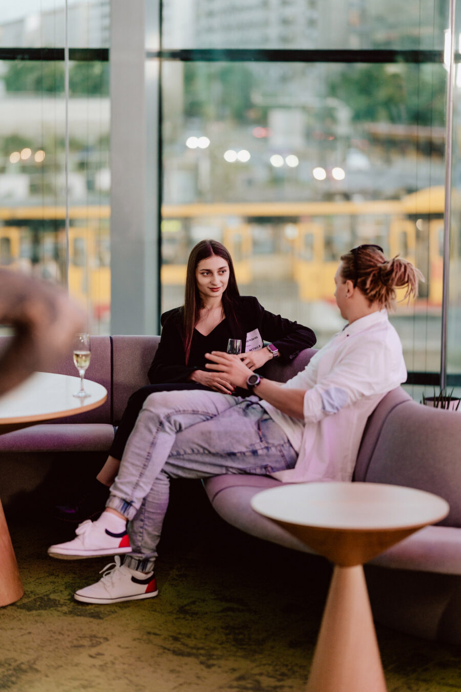 fotograf startupow rotunda reaktorX 93 Zwei Personen sitzen auf einem modernen Sofa in einem gläsernen Wohnzimmer und führen ein Gespräch. Auf dem Tisch neben ihnen steht ein Glas Wein. Draußen vor dem Fenster sehen Sie gelbe Straßenbahnen und städtische Gebäude.