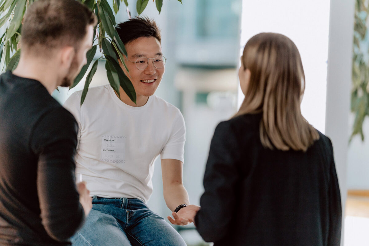 fotograf startupow rotunda reaktorX 94 Drei Personen unterhalten sich in einem Raum. Ein Mann mit weißem T-Shirt und Brille spricht und lächelt, während ein Mann und eine Frau, beide in dunkler Kleidung, aufmerksam zuhören. Über ihnen hängen grüne Blätter.