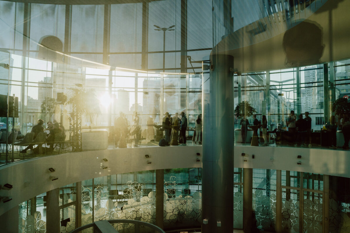 fotograf startupow rotunda reaktorX 95 Das Sonnenlicht strömt durch große Glasfenster in ein modernes, rundes Gebäude mit mehreren Stockwerken. Die Silhouetten der Menschen sind verstreut, einige sitzen, andere stehen und schaffen eine geschäftige, urbane Atmosphäre.