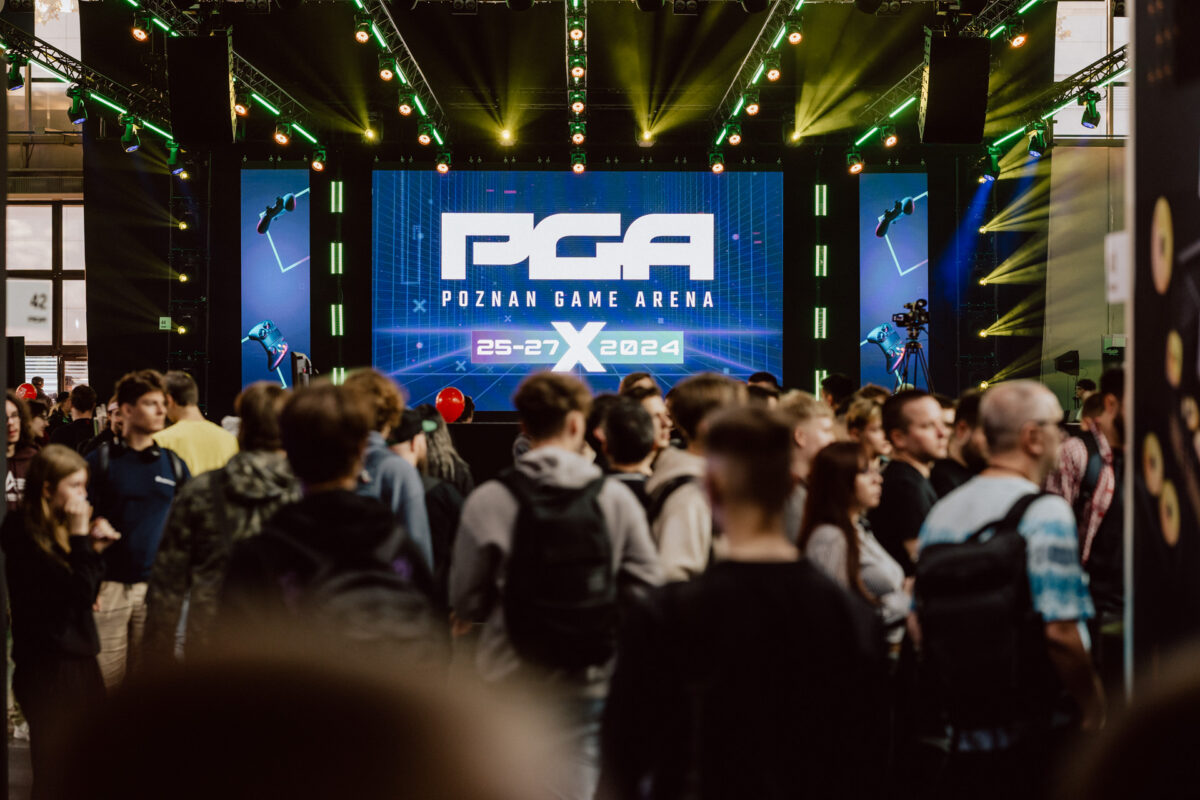 Messefotograf poznań - Eine große Menschenmenge versammelt sich vor einer Bühne mit einem großen Bildschirm, auf dem "PGA Poznań Game Arena 25-27 X 2024" angezeigt wird, während der Gaming-Veranstaltung, mit grünen Lichtern und Gaming-Dekorationen über dem Kopf.