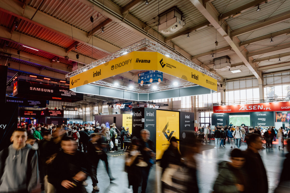 Eine belebte Messehalle mit einem Gewimmel von Menschen, die an Ständen und Auslagen von Unternehmen wie Intel, Samsung und Alsen vorbeiziehen, unter einer hohen Decke mit hellen Lichtern und einem großen gelben Banner.